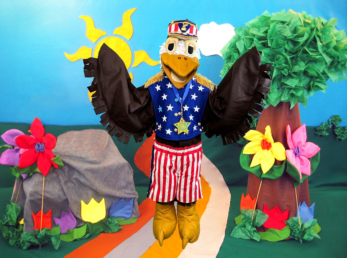 Scout Regal Eagle, a costumed eagle character in a red, white and blue patriotic outfit, standing on a garden path with a sun and trees in the background